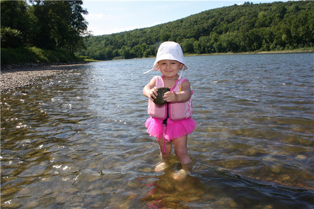 First Place Winner - So Proud - Photographer: Erika Bruckner. Toddler age girl standing in shallow creek water holding a rock, looking very proud of herself.