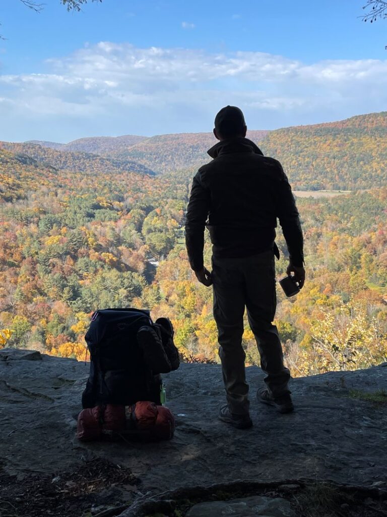 A man with a backpack looks at a view of mountains and fall foliage