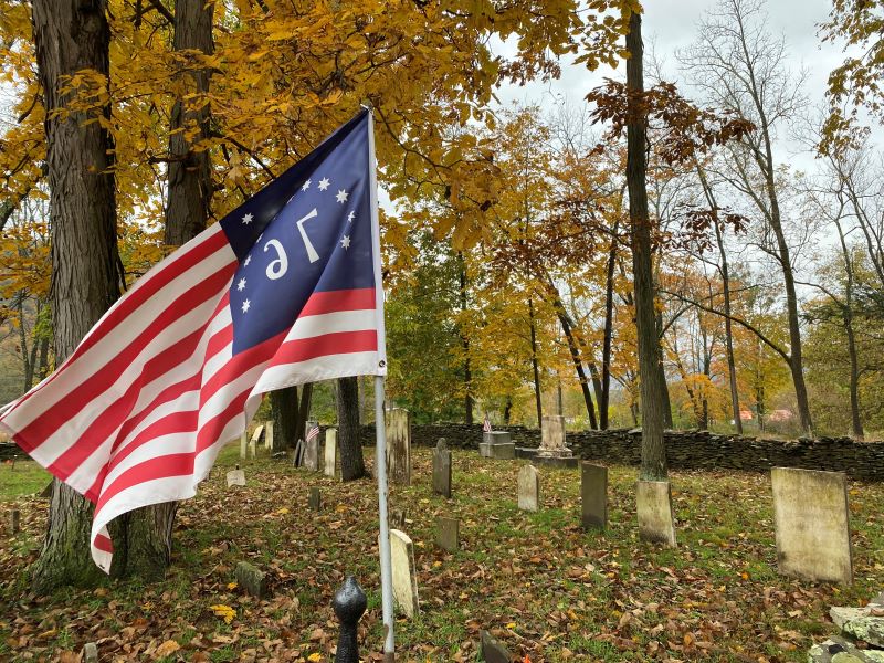 A colonial flag at Marcy Cemetery with headstones and fall foliage in the background